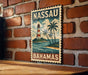 A vintage-style metal sign with the text "NASSAU BAHAMAS" and an illustration of a lighthouse and palm trees, mounted on a brick wall.