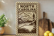 a decorative sign that reads "NORTH CAROLINA" with a lighthouse and mountains in the background. The sign is placed on a shelf next to a potted plant and a cutting board, along with some lemons.