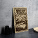 a wooden sign with the text "NORTH CAROLINA" and a lighthouse illustration, placed on a countertop next to some black camera lenses.