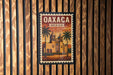 A vintage-style poster of Oaxaca, Mexico, featuring a building with a dome and palm trees, is displayed on a wooden wall.