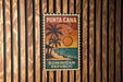 A vintage-style poster of Puna Cana, the Dominican Republic, featuring a palm tree and sunset, is displayed on a wooden wall.