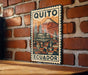 A metal sign with a vintage-style illustration of Quito, Ecuador, featuring a mountain and a cityscape.