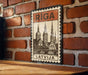 A vintage-style metal sign with a brick wall backdrop, featuring a drawing of a city skyline and the word "Riga" prominently displayed.