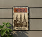 A vintage-style postage stamp featuring the cityscape of Riga, Latvia, with the word "Riga" prominently displayed at the top.
