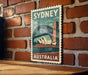A vintage-style poster of Sydney, Australia, featuring the iconic Sydney Opera House and the Sydney Harbour Bridge, is displayed on a brick wall.