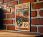 A vintage-style postage stamp from Tennessee, featuring a mountain landscape and the state's name, is displayed on a brick wall.