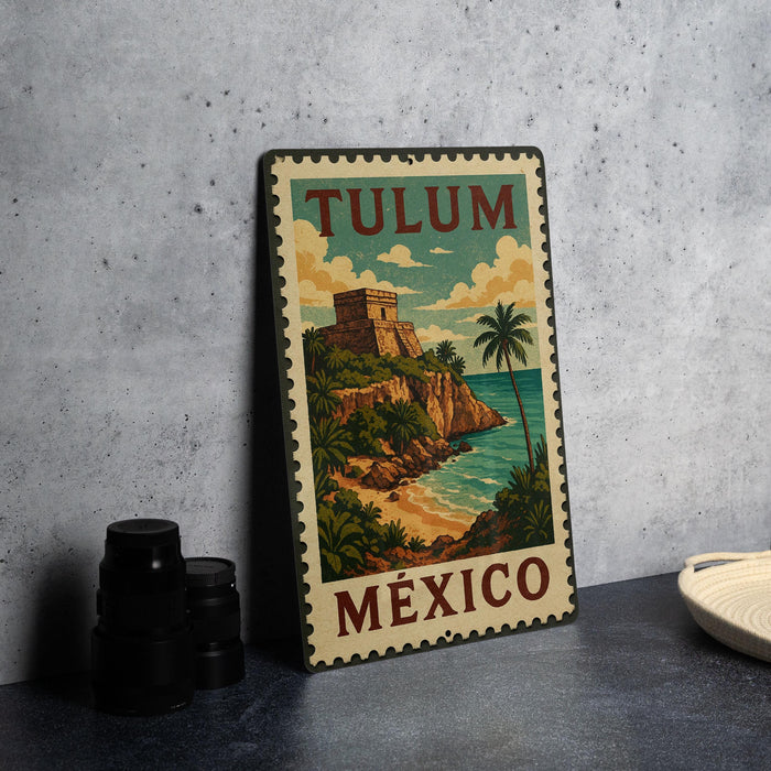 A vintage-style metal sign with the text "TULUM MEXICO" and an illustration of a tropical beach and castle, placed on a gray countertop next to some black camera lenses.