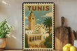 A vintage-style postage stamp depicting the city of Tunis, Tunisia, with a tall minaret and palm trees, is displayed on a shelf next to kitchen utensils and a potted plant.