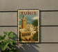 A vintage-style postage stamp depicting a cityscape of Tunis, Tunisia, with a tall tower and palm trees.
