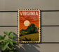 A vintage-style postage stamp depicting a rural landscape with a large sun in the sky, mounted on a wall next to a plant.