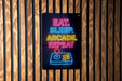 A neon sign with the text "EAT. SLEEP. arcADE. REPEAT" in neon colors is displayed on a wooden wall.
