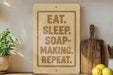 a wooden sign with the text "EAT. SLEEP. SOAP-MAKING. REPEAT." on it, placed on a wooden shelf next to a potted plant and kitchen utensils.