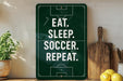 a green sign with white text that reads "EAT. SLEEP. SOCCER. REPEAT." The sign is placed on a shelf next to a wooden cutting board and a potted plant.