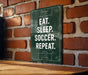 A green sign with white text is mounted on a brick wall, displaying the message "EAT. SLEEP. SOCCER. REPEAT."