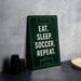 a green sign with white text that reads "EAT. SLEEP. SOCCER. REPEAT." The sign is placed on a gray countertop next to a stack of black containers and a white bowl.
