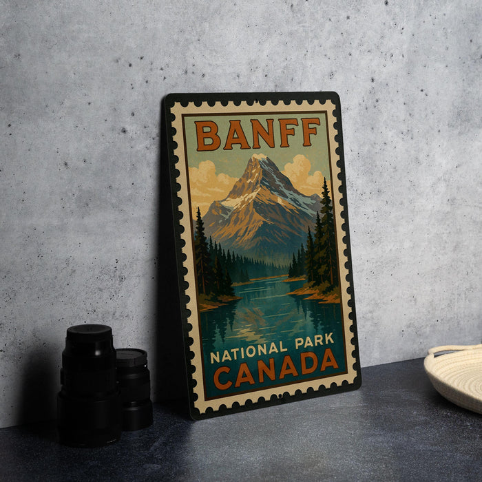 A vintage-style metal stamp depicting a mountain landscape with a lake, surrounded by pine trees, is displayed on a gray countertop next to some black camera lenses.