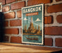 A vintage-style poster of Bangkok, Thailand, featuring a temple and a boat on the water, mounted on a brick wall.