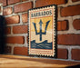 A vintage-style metal sign with the word "BARBADOS" and a trident symbol is displayed on a brick wall.