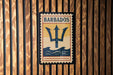 A vintage-style postage stamp with the word "BARBADOS" printed on it, featuring a trident symbol, is displayed on a wooden wall.