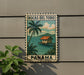 A vintage-style metal sign with a tropical island scene and palm trees, featuring the text "BOCAS DEL TORO PANAMA" and a postage stamp-like border.