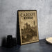 A metal sign with the text "CARDIFF WALES" and an illustration of a castle and bridge, placed on a table next to some black camera lenses.