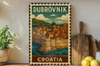 A vintage postage stamp depicting Dubrovnik, Croatia, with a cityscape and a castle, is displayed on a shelf next to a wooden cutting board and a potted plant.