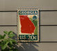 A U.S. postage stamp from Georgia, featuring a red state outline and a green field with a tree, mounted on a wall.