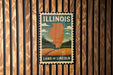 A vintage-style postage stamp depicting the state of Illinois, with a landscape of fields and forests in the background.