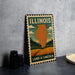 A vintage-style metal sign with the text "ILLINOIS" and "LAND OF LINCOLN" printed on it, depicting a map of Illinois with a landscape in the background.