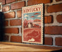 A vintage-style metal sign with a stamp-like border, depicting a rural landscape with a house and trees, mounted on a brick wall.