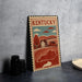 A vintage-style metal sign with a stamp-like border, depicting a rural landscape with a house and trees, is displayed on a gray countertop next to some black camera lenses.