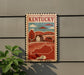 A vintage-style sign depicting a rural landscape with a house and trees, mounted on a wall next to a plant.