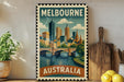 A vintage-style metal stamp depicting the Melbourne skyline, with a bridge and water in the foreground, is displayed on a shelf next to a wooden cutting board and some lemons.