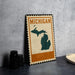 A metal sign with the word "MICHIGAN" on it, featuring a map of Michigan, is displayed on a table next to some black camera lenses.