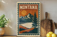 A vintage-style poster of Montana mountains and a lake, featuring a pine tree and a sunset, is displayed on a wall next to a wooden cutting board and a potted plant.