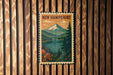 A vintage-style postage stamp depicting a scenic landscape with a mountain, lake, and pine trees, hanging on a wooden wall.