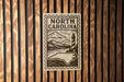 A metal sign with the text "NORTH CAROLINA" and an illustration of a lighthouse and mountains, mounted on a wooden wall.