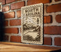 A decorative metal sign with the text "NORTH CAROLINA" and an illustration of a lighthouse and mountains, mounted on a brick wall.