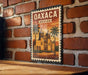 A vintage-style poster of Oaxaca, Mexico, featuring a brick wall and palm trees, is displayed on a wooden surface.