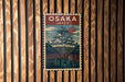A vintage-style poster of Osaka, Japan, featuring a castle-like structure and a bridge, is displayed on a wooden wall.