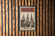A vintage-style postage stamp from Latvia featuring a cityscape with a bridge and a church, mounted on a wooden wall.