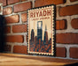 A vintage-style metal sign with the text "RIYADH SAUDI ARABIA" and an illustration of the skyline of Riyadh, Saudi Arabia, mounted on a brick wall.