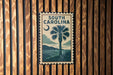 A framed poster of a palm tree and a sunset is displayed on a wooden wall.