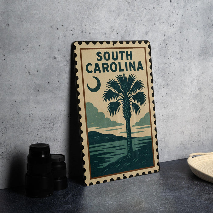 A metal sign with the text "SOUTH CAROLINA" and a palm tree illustration, placed on a gray countertop next to some black camera lenses.