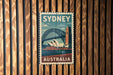 A vintage-style poster of Sydney, Australia, featuring the iconic Sydney Opera House and the Sydney Harbour Bridge, is displayed on a wooden wall.