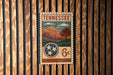 A vintage-style postage stamp from Tennessee, featuring a mountain landscape and a tree, is displayed on a wooden wall.