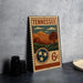 A vintage-style metal stamp from Tennessee featuring a landscape scene with mountains, trees, and a tree with three stars.