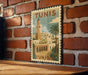A vintage-style metal sign with the text "TUNIS" and an illustration of a cityscape with a tall tower, palm trees, and a mosque, mounted on a brick wall.