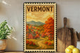 A framed vintage-style poster of a rural landscape in Vermont, featuring a red barn, a winding road, and colorful autumn foliage.