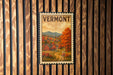 A vintage Vermont postage stamp is displayed on a wooden wall, featuring a scenic autumn landscape with a red barn and a winding road.